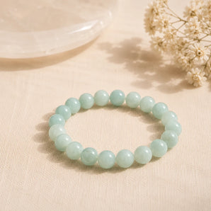 A beaded stretch bracelet made of smooth, pale mint-green round Amazonite crystal beads, displayed on a textured beige fabric surface with dried flowers and a soft-focus glass bowl in the background.