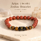 A beaded Aries Zodiac Bracelet made of polished red jasper and tiger eye, featuring a central black cube spacer with a gold Aries symbol. The bracelet is displayed on a light stone slab with decorative dried flowers and text overlays.