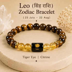 A beaded astrology bracelet for Leo zodiac, featuring brown tiger's eye and clear citrine beads with a central black cube charm engraved with a gold lion and the word 'Leo'. The bracelet is displayed on a beige stone slab with decorative crystals in the background.