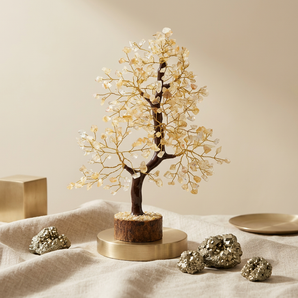A decorative crystal tree figurine with a dark brown trunk and branches adorned with clear and pale gold gemstones, displayed on a round gold-toned pedestal. The tree sits on draped beige fabric with metallic gold crystals and a small gold dish nearby against a warm beige background.