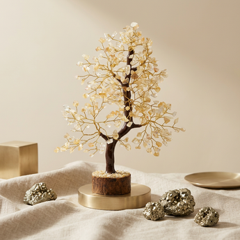 A decorative crystal tree figurine with a dark brown trunk and branches adorned with clear and pale gold gemstones, displayed on a round gold-toned pedestal. The tree sits on draped beige fabric with metallic gold crystals and a small gold dish nearby against a warm beige background.