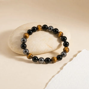 Triple Protection bracelet with black obsidian, golden tiger eye and grey hematite beads resting on a cream stone – World of Oorja