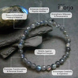 Gray beaded bracelet with text highlighting its benefits on a stone background, branded 'World of Orja'.