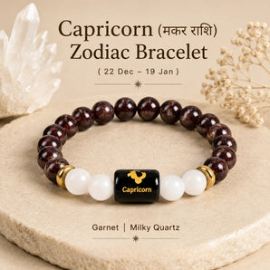 A beaded bracelet displayed on a stone slab with decorative crystals and dried flowers. The bracelet has dark reddish-brown garnet beads, white quartz beads, gold spacer rings, and a central black cube with a gold Capricorn zodiac symbol.