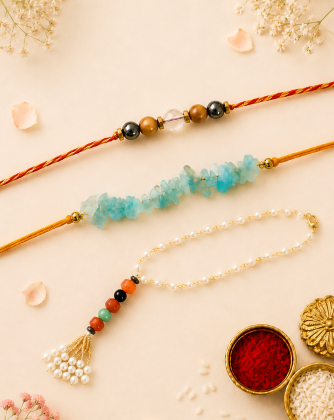 Three crystal rakhi bracelets arranged on a light beige background with dried flowers and small bowls of red and white powder. One bracelet has a coral-red cord, one has a tan cord with turquoise-blue stone beads, and one has a white pearl cord with gold tassel and multicolor beads.