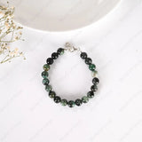 Beaded bracelet with green and black stones on a white surface with a small plant.