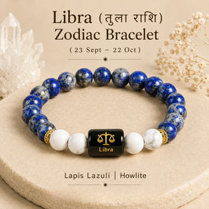A Libra zodiac bracelet made of blue and white marbled beads with a central black cube featuring a gold Libra symbol and the word 'Libra'. The bracelet is displayed on a beige stone surface with decorative crystals and dried flowers in the background.