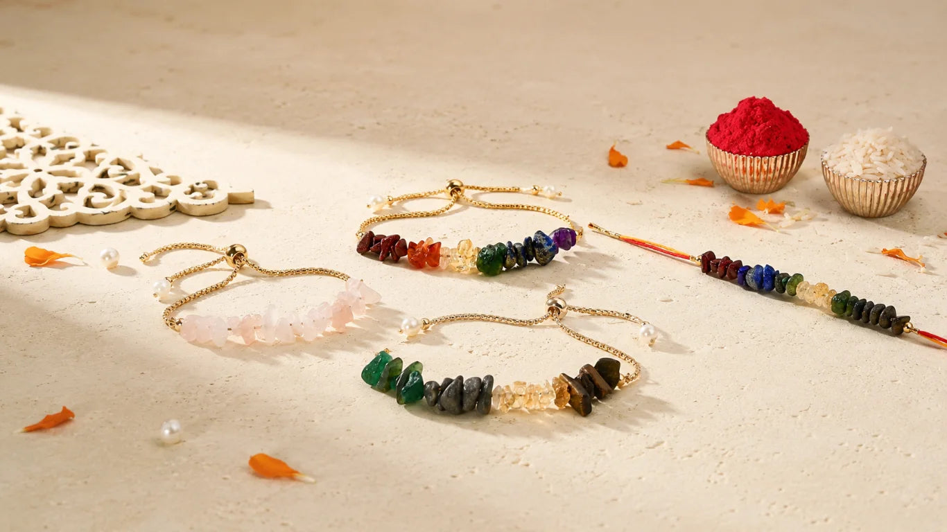 Four gold-toned crystal rakhi arranged on a light beige surface with decorative props, including small bowls of red and white powder and scattered orange petals. The bracelets feature multicolored stone beads and one pale pink crystal pendant.