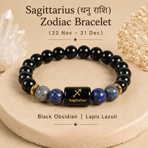 A Sagittarius zodiac bracelet made of black obsidian and lapis lazuli beads, displayed on a beige stone platform with decorative crystals and dried flowers. The central bead is black with gold lettering and the Sagittarius zodiac symbol.