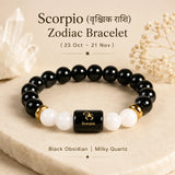 A Scorpio Zodiac bracelet made of glossy black obsidian beads, white milky quartz beads, and gold spacer rings, displayed on a beige stone slab with decorative crystals and dried flowers. The central bead is black with a gold Scorpio symbol.