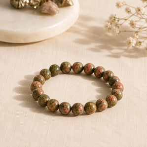 A beaded stretch bracelet made of polished round unakite crystal beads in earthy green, brown, and pink tones, arranged on a light beige fabric surface with soft decorative props in the background.