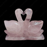 Rose quartz crystal sculpture of two swans forming a heart shape on a black background.