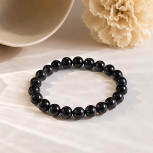 Black Obsidian bracelet with glossy jet-black crystal beads on cream draped fabric beside a ceramic bowl – World of Oorja