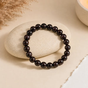 Garnet bracelet with deep wine-red glossy beads on a smooth cream stone with dried pampas and a lit candle – World of Oorja