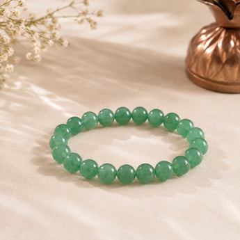 Jade bracelet with mottled green beads on soft cream fabric beside a copper kalash and white dried flowers – World of Oorja