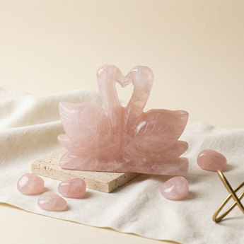 A pink rose quartz swan figurine carved with a heart-shaped nest, displayed on a small stone block with several loose rose quartz crystals on a cream fabric surface.