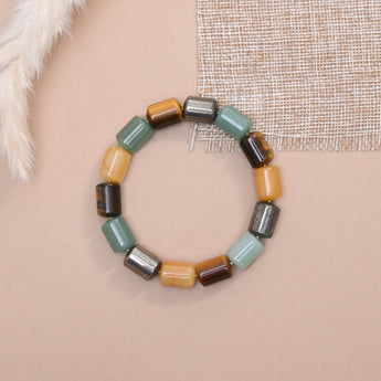 A beaded stretch bracelet made of cylindrical stone beads of green aventurine, pyrite, tiger eye, citrine  arranged in a circle on a beige background with decorative fabric and faux fur props.