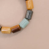 A close-up of a beaded strand with cylindrical beads of green aventurine, pyrite, tiger eye, citrine set against a solid pale pink background.