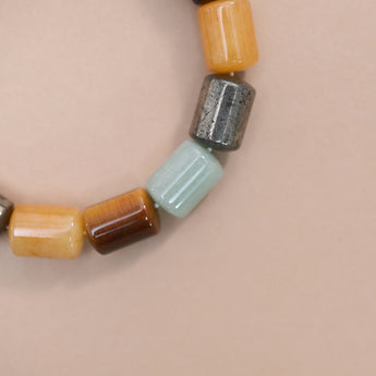 A close-up of a beaded strand with cylindrical beads of green aventurine, pyrite, tiger eye, citrine set against a solid pale pink background.