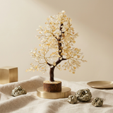 A decorative crystal tree figurine with a dark brown trunk and branches adorned with clear and pale gold gemstones, displayed on a round gold-toned pedestal. The tree sits on draped beige fabric with metallic gold crystals and a small gold dish nearby against a warm beige background.