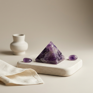 A purple amethyst crystal pyramid displayed on a light beige stone slab, with two small oval amethyst stones beside it and a small white ceramic vase in the background on a warm beige surface.