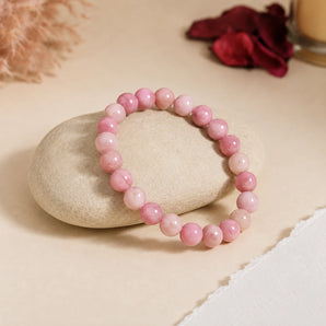 Rhodochrosite bracelet with blush to deep rose banded beads draped over a beige river stone with red rose petals – World of Oorja