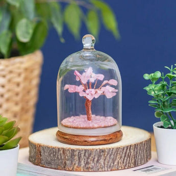 Decorative tree with rose quartz blossoms under a glass dome on a wooden base, surrounded by plants.